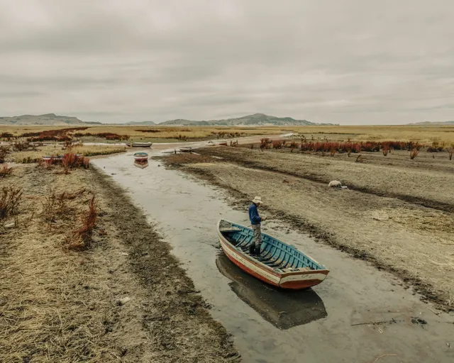Vista elevada de un pequeño bote varado o navegando a duras penas en un hilo mínimo de agua en medio del barro y la pampa árida.