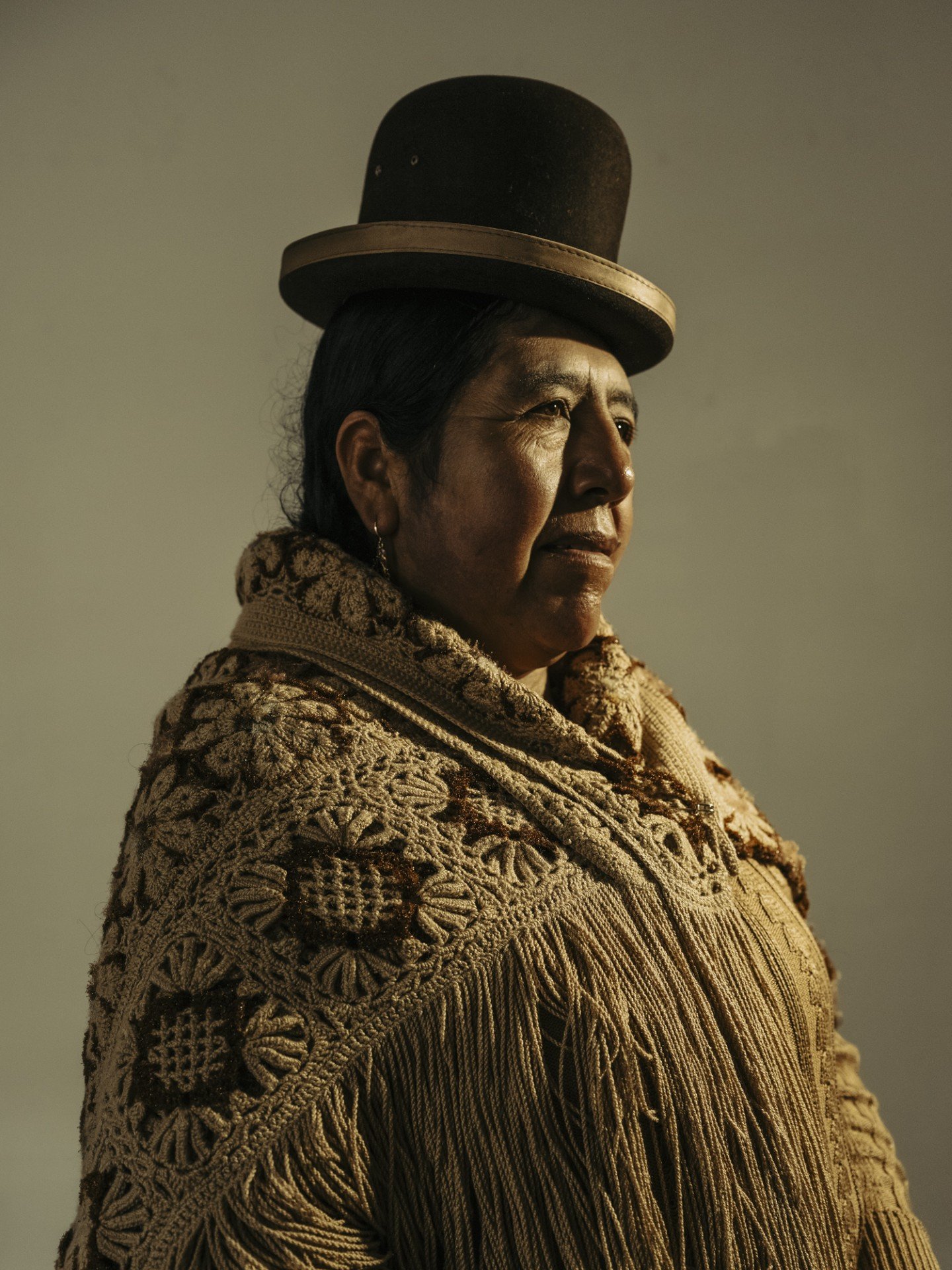 Retrato formal de estudio/iluminación dramática de una mujer andina vistiendo un sombrero de copa (bombín) y mantilla gruesa, con un perfil fuerte y digno frente a un fondo oscuro.
